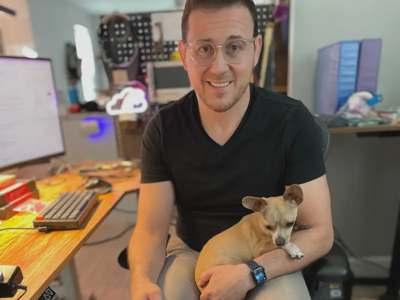 A man in a black v-neck shirt sitting at a desk looking into the camera smiling with a tiny chihuachua in his lap