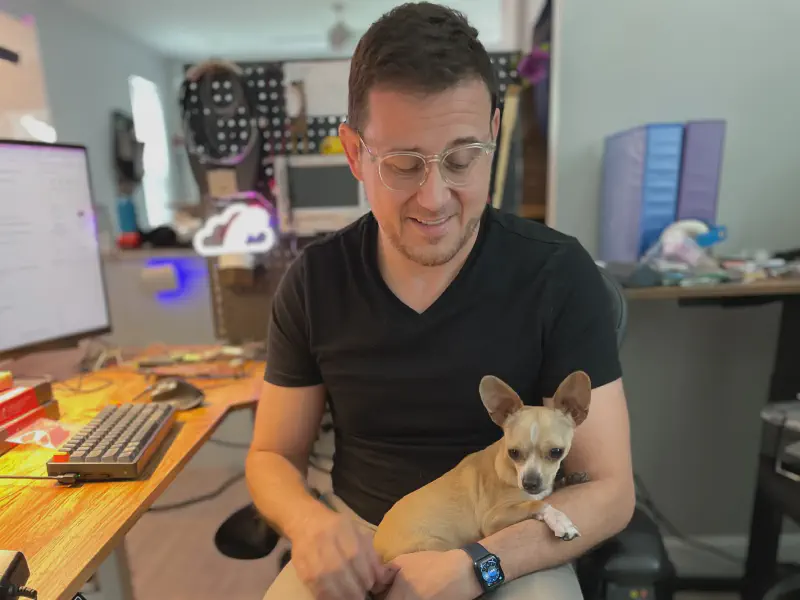 A man in a black v-neck shirt sitting at a desk looking down at a tiny chihuachua in his lap