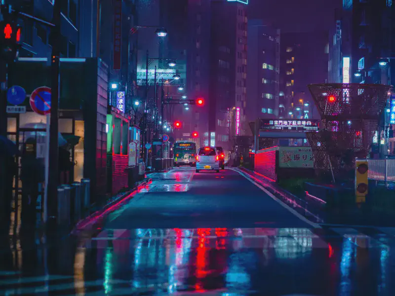 A view down a dark city steet at night with tail lights and the glow of neon lights in the distance